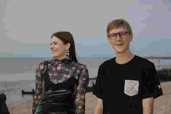 Alev and Jas are on the beach, leanging on a wooden fence the sea and pebble beach visible behind them. Alev looks to the left and laughs, wearing a clue shimmering dress atop a flowery long armed mesh top with big earrings, three hearts dangling on top of each other and a pearl. Jas laughs too and faces the camera with a black short sleeved shirt with a pocket with also flowers on it.